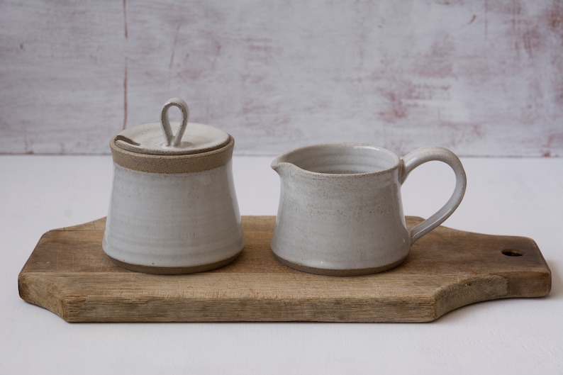 Op de afbeelding: Een witte keramische roomkan en suikerpot set met een deksel op een houten snijplank. De roomkan heeft een handvat en de suikerpot heeft een deksel met een klein handvat.