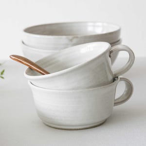 May include: A stack of two white ceramic mugs with handles, one holding a wooden spoon. Several white bowls are stacked in the background. The mugs and bowls have a rustic, handmade appearance, suggesting a kitchenware theme.