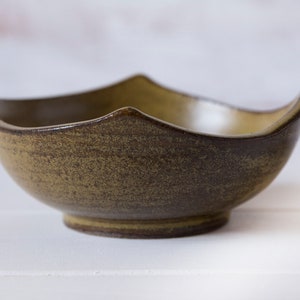 Può includere: Una ciotola in ceramica fatta a mano con un bordo ondulato unico. La ciotola ha una glassa macchiata marrone terroso e verde oliva. La ciotola è rotonda con una piccola base circolare. La ciotola viene probabilmente utilizzata per servire cibo.