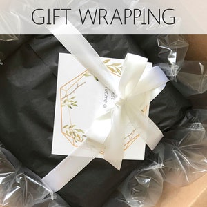 Op de afbeelding: Een cadeau verpakt in zwart tissuepapier met een wit lint en een kaart met de tekst "De beste tijd is een leven lang" in een gouden geometrisch frame.