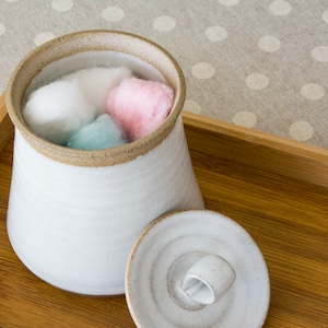Op de afbeelding: Een witte keramische pot met een deksel, gevuld met wattenbolletjes in roze, blauw en wit. De pot staat op een houten dienblad met een lichtbruin stippenpatroon.