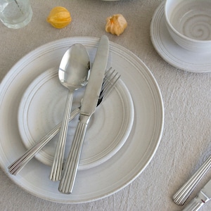 May include: A white ceramic dinner plate with a smaller white ceramic plate inside. A silver fork, knife, and spoon are placed on top of the plates.