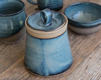 May include: A blue and white ceramic jar with a lid. The jar has a handle on the lid and a light blue glaze. The jar is sitting on a wooden surface.