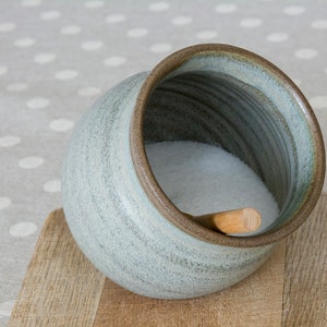 Puede incluir: Un salero de cerámica gris con una cuchara de madera en su interior, lleno de sal blanca. El salero está sobre una tabla de cortar de madera.