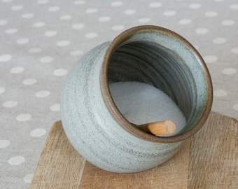 Peut inclure: Une salière en céramique grise avec une cuillère en bois à l'intérieur, remplie de sel blanc. La salière est posée sur une planche à découper en bois.