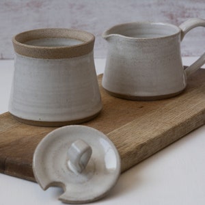 Op de afbeelding: Een witte keramische suikerpot met een deksel en een bijpassende roomkan. De suikerpot en de roomkan staan op een houten snijplank.