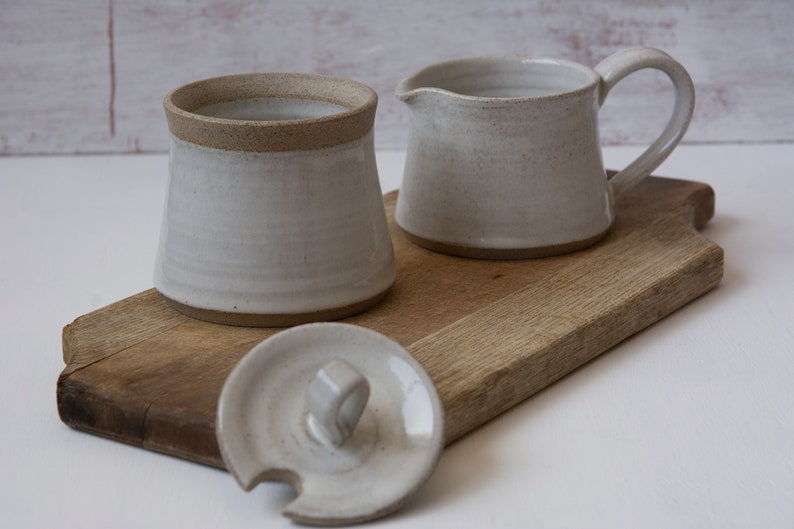 Op de afbeelding: Een witte keramische suikerpot met een deksel en een bijpassende roomkan. De suikerpot en de roomkan staan op een houten snijplank.