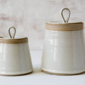 Könnte beinhalten: Zwei weiße Keramikdosen mit Deckeln und braunem Rand. Die Dosen haben ein schlichtes, elegantes Design und eignen sich perfekt zum Aufbewahren von Zucker, Mehl oder anderen Küchenzutaten.