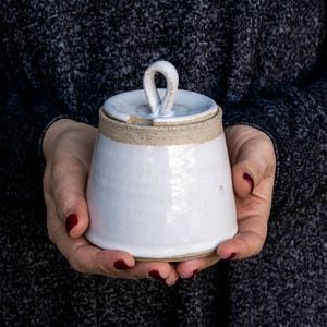 Op de afbeelding: Witte keramische pot met een deksel met een lusvormig handvat. De pot heeft een getextureerd, licht ruw oppervlak.