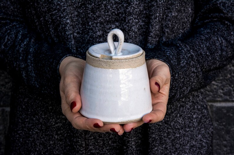 Op de afbeelding: Witte keramische pot met een deksel met een lusvormig handvat. De pot heeft een getextureerd, licht ruw oppervlak.