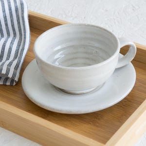 May include: A white ceramic teacup and saucer set on a wooden tray. The cup has a textured, slightly rippled surface and a rounded handle.