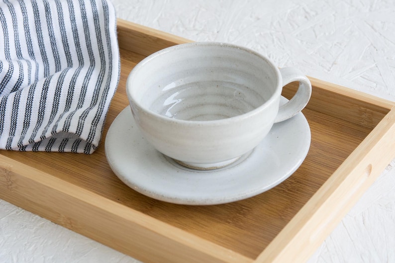 May include: A white ceramic teacup and saucer set on a wooden tray. The cup has a textured, slightly rippled surface and a rounded handle.
