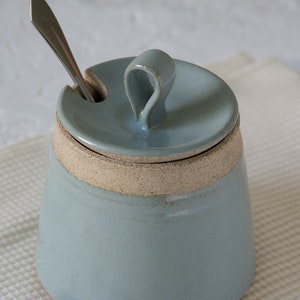 May include: A light blue ceramic sugar jar with a lid and a small spoon. The jar has a white band around the middle and a textured finish.