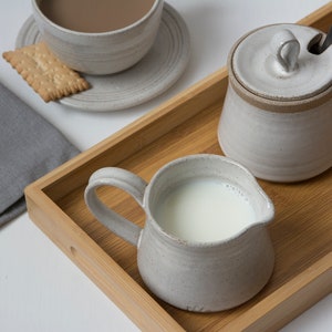 May include: A wooden tray with a white ceramic creamer, sugar bowl with lid, and a saucer with a cup and a cookie. The tray is made of light brown wood and has a natural finish.