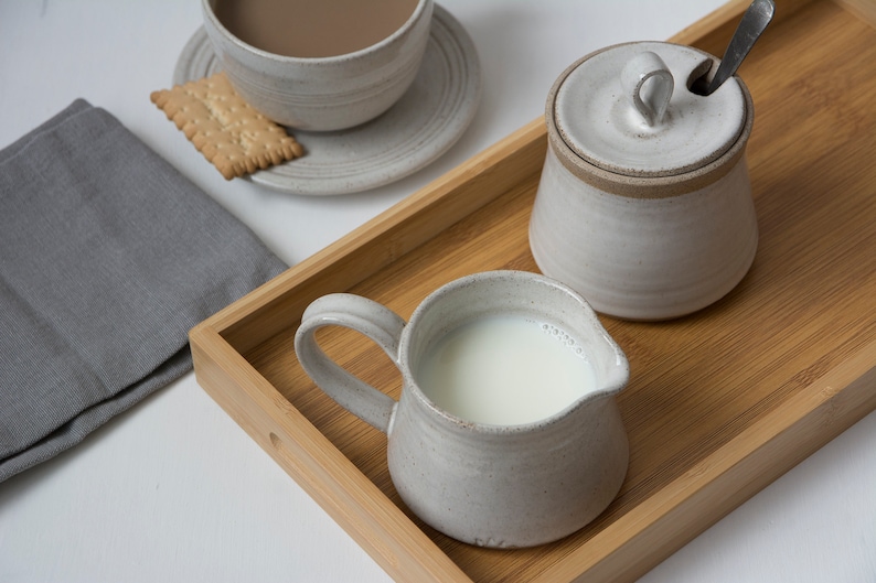 Op de afbeelding: Een houten dienblad met een witte keramische kan, een suikerpot met deksel en een schoteltje met een kopje en een koekje. Het dienblad is gemaakt van lichtbruin hout en heeft een natuurlijke afwerking.