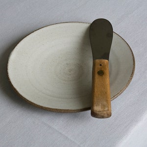 Puede incluir: Un plato de cer&aacute;mica blanco con un borde marr&oacute;n y un cuchillo de mantequilla de metal con mango de madera.