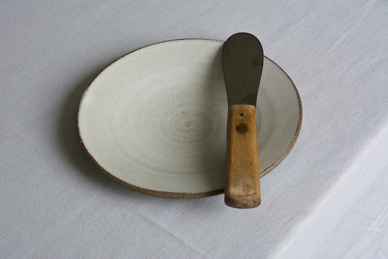 Puede incluir: Un plato de cer&aacute;mica blanco con un borde marr&oacute;n y un cuchillo de mantequilla de metal con mango de madera.