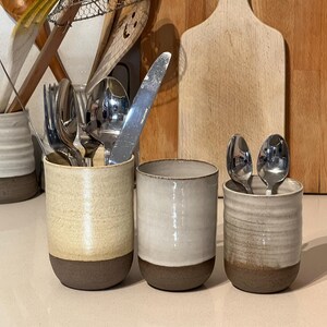 Könnte beinhalten: Drei Keramik-Utensilienhalter in Creme, Beige und Weiß, jeweils mit einem dunkelbraunen Boden. Die Halter enthalten Besteck, darunter Gabeln, Löffel und ein Messer. Schneidebretter aus Holz und Küchenutensilien befinden sich im Hintergrund.