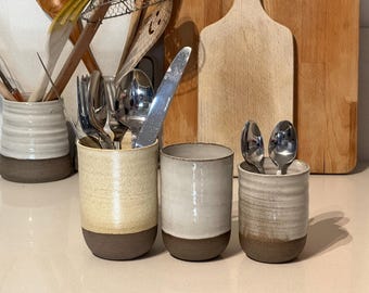 May include: Three ceramic utensil holders in cream, beige, and white, each with a dark brown base. The holders contain silverware, including forks, spoons, and a knife. Wooden cutting boards and kitchen utensils are in the background.