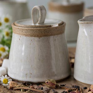 Op de afbeelding: Een set witte keramische keukenartikelen met een bruine rand. De set bevat een roomkan, een suikerpot met deksel en een kan. De items zijn gerangschikt op een houten oppervlak met gedroogde bloemen en specerijen.
