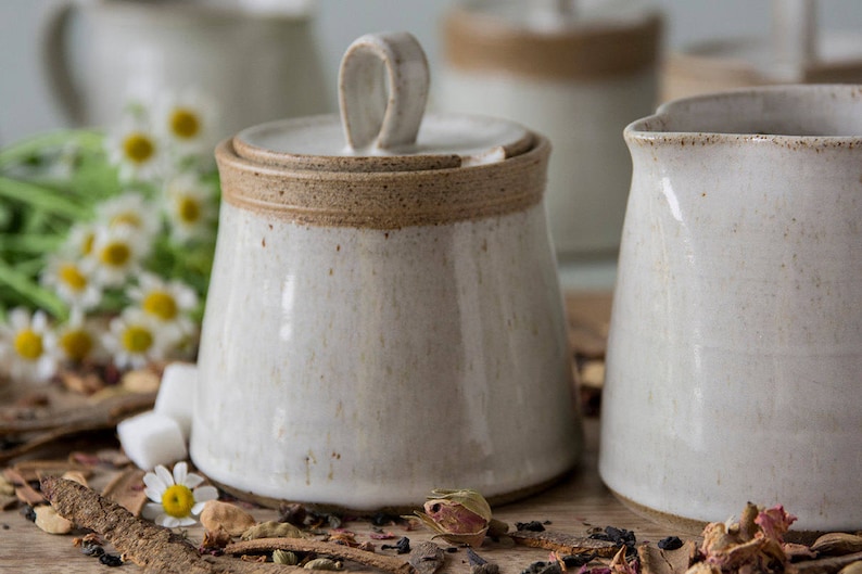 Op de afbeelding: Een set witte keramische keukenartikelen met een bruine rand. De set bevat een roomkan, een suikerpot met deksel en een kan. De items zijn gerangschikt op een houten oppervlak met gedroogde bloemen en specerijen.