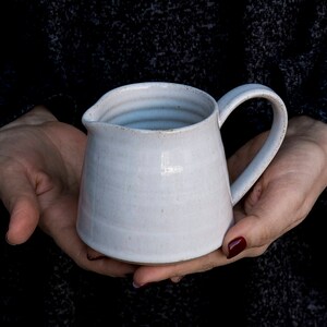Op de afbeelding: Een witte keramische kan met een rond lichaam en een handvat. De kan heeft een glad, licht getextureerd oppervlak.