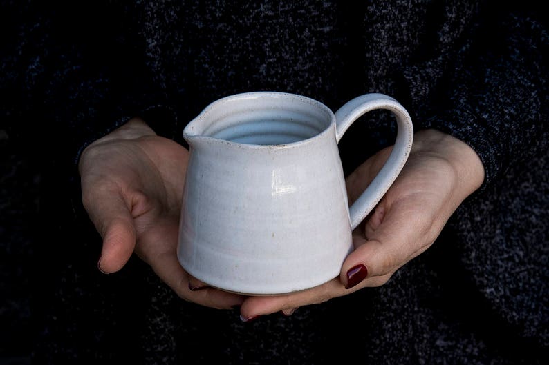 Op de afbeelding: Een witte keramische kan met een rond lichaam en een handvat. De kan heeft een glad, licht getextureerd oppervlak.