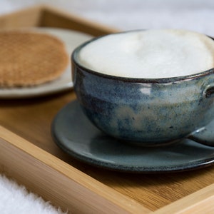 May include: A blue ceramic mug filled with a creamy cappuccino sits on a blue saucer on a wooden tray. A round, brown cookie is on a white saucer next to the mug.