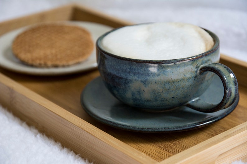 May include: A blue ceramic mug filled with a creamy cappuccino sits on a blue saucer on a wooden tray. A round, brown cookie is on a white saucer next to the mug.