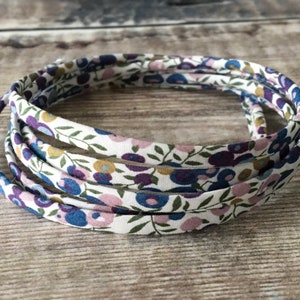 May include: A close-up of a white fabric trim with a floral pattern in shades of purple, blue, and pink. The trim is coiled into a circle and is approximately 2.5 cm wide.