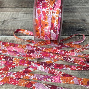 May include: A roll of floral ribbon in a clear container with loose ribbon strands. The ribbon features a repeating pattern of small flowers in shades of pink, orange, and red. The ribbon is approximately 1.3 cm wide.