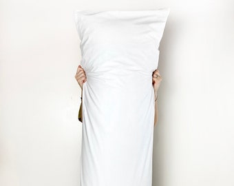May include: A white rectangular pillow being held up by a person in front of a white wall. The pillow is large and covers the person's body from the shoulders down.