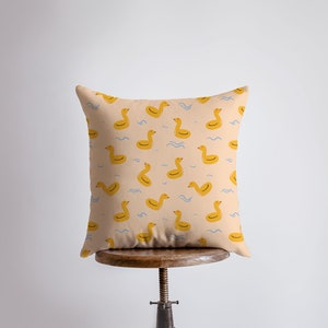 May include: Square cushion featuring a pattern of yellow inflatable ducks on a pale peach background. Blue wavy lines are between the ducks. The cushion is displayed on a wooden stool.