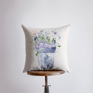 Puede incluir: Una almohada blanca con una ilustración de acuarela de una maceta azul con flores moradas y mariposas.