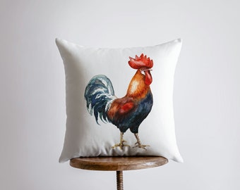 May include: White cushion with a watercolour illustration of a rooster with a red comb and wattle, blue and brown feathers, and a white background.