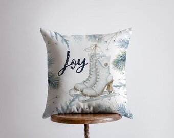 May include: White throw pillow with a watercolor design of ice skates and pine branches. The pillow has the word "Joy" written in black script.