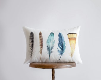 Puede incluir: Una almohada rectangular blanca con cinco plumas de acuarela impresas en ella. Las plumas son en tonos de azul, marrón y amarillo. La almohada está sentada en un taburete de madera.