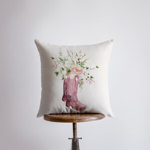Puede incluir: Una almohada blanca con un diseño floral rosa que presenta un ramo de flores en un par de botas de lluvia rosas.