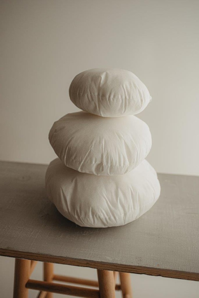 May include: Three white, round cushions stacked. The cushions have a textured surface and are placed on a grey surface. The background is a neutral colour. The cushions are approximately 30 cm in diameter.