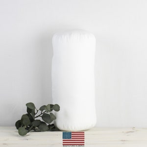 Puede incluir: Almohada cilíndrica blanca con una funda de tela lisa. La almohada está sobre una superficie de madera clara con una rama de hojas verdes delante. El texto "MANUFACTURED IN AMERICA" está impreso debajo de una pequeña bandera estadounidense.