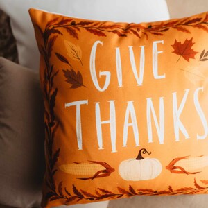 Pu&ograve; includere: Cuscino arancione con il testo "Give Thanks" in lettere bianche. Il cuscino ha un tema autunnale con illustrazioni di foglie, mais e una zucca.