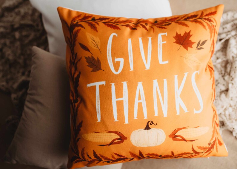 Pu&ograve; includere: Cuscino arancione con il testo "Give Thanks" in lettere bianche. Il cuscino ha un tema autunnale con illustrazioni di foglie, mais e una zucca.