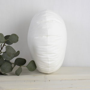 May include: A white, oval cushion with a soft, textured surface. The cushion is standing upright, highlighting its shape and design. The background is a neutral white, with a sprig of green leaves in the lower left corner.