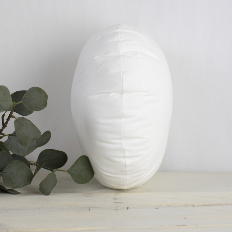 May include: A white, oval cushion with a soft, textured surface. The cushion is standing upright, highlighting its shape and design. The background is a neutral white, with a sprig of green leaves in the lower left corner.