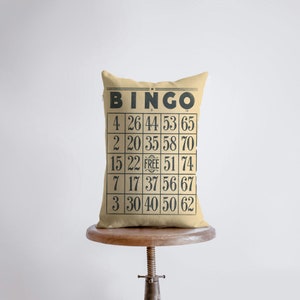 May include: A beige pillow with a black and white bingo card design. The card has the word "BINGO" at the top and numbers in each square. The free space is marked with the word "FREE".
