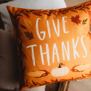 Pu&ograve; includere: Cuscino arancione con il testo "GIVE THANKS" in lettere bianche. Il cuscino &egrave; decorato con foglie autunnali, una zucca e mais.
