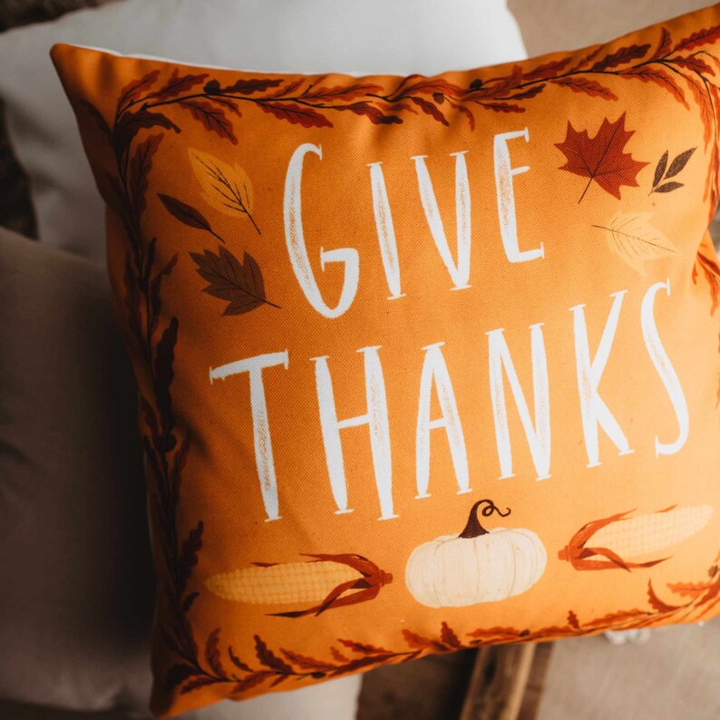 Pu&ograve; includere: Cuscino arancione con il testo "GIVE THANKS" in lettere bianche. Il cuscino &egrave; decorato con foglie autunnali, una zucca e mais.