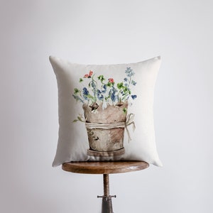 Puede incluir: Una almohada blanca con una ilustración de acuarela de una maceta de terracota llena de flores azules y verdes y mariquitas rojas.