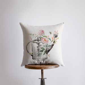Puede incluir: Una almohada blanca con una ilustración de acuarela de una regadera llena de flores rosas y blancas. La regadera es gris y tiene un pico y un asa. Hay abejas volando alrededor de las flores.