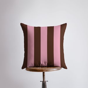 May include: Square decorative pillow with a brown border and alternating vertical stripes of brown and light pink. The pillow is resting on a wooden stool against a white background. The pillow is a home decor item.
